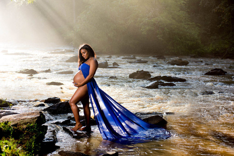 A pregnant woman cradling her pregnant belly for her magazine style maternity photoshoot while wearing a flowing navy blue fabric while standing on a rock on flowing river that is behind her, Moreland Photography, Atlanta maternity photography, magazine style maternity photo, maternity portrait,  Atlanta maternity portrait, river, river rocks, blue fabric, blue flowing fabric, water, flowing river, atlanta river