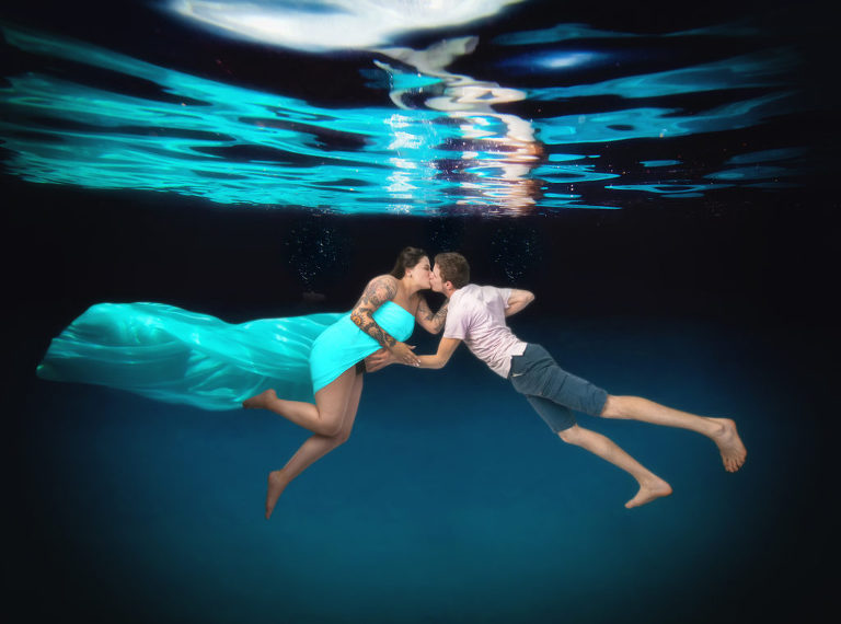 A couple kissing for their underwater magazine style maternity shoot while the man cradles the woman's pregnancy belly and the woman is wearing a bright teal fabric garment while the man wears a white pink shirt with navy grey shorts, Moreland Photography, Atlanta maternity photography, magazine style maternity photo, maternity portrait,  Atlanta maternity portrait, underwater photography, underwater maternity portrait, underwater portrait, underwater maternity photography, underwater photography, atlanta underwater portrait