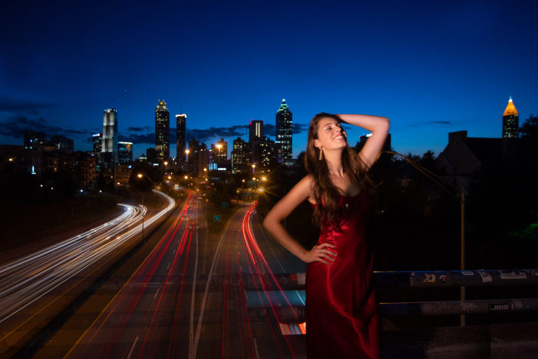 A girl smiling for her senior portrait while also doing a dynamic pose of one hand on her hip with her other hand on her head as she wear a long red silky dress with the background of the skyline from Atlanta at night, Moreland Photography, Atlanta, Atlanta Georgia, Skyline, Senior, Senior Portrait, Portraiture, Red Dress, Senior picture, Red Silk dress, Silk Dress, Skyline at night, Night time, Creative Lighting, Dusk Lighting, City, Vibrant city