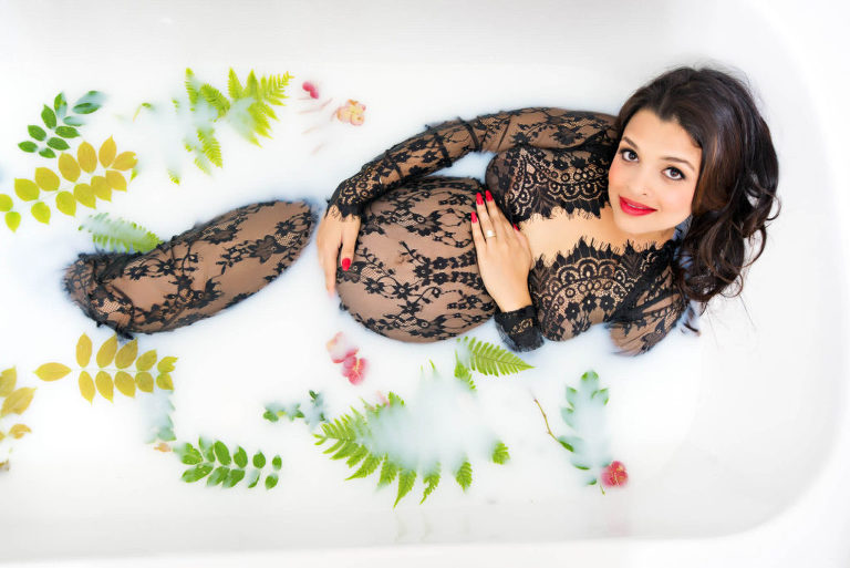 A pregnant woman cradling her pregnant belly while wearing a black lacy long outfit with long brown hair, wearing red lipstick and red nails while laying in water filled with leaves, Moreland Photography, Atlanta maternity photography, magazine style maternity photo, maternity portrait,  Atlanta maternity portrait, red nails, red lipstick, pregnant belly, water with leaves, creamy water with leaves and greenery, greenery, black lace outfit