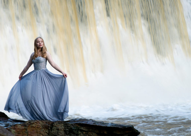 A girl who is a senior smiling and posing for her senior portrait while wearing a long silver white dress that a sparkly top as she holds out the skirt of the dress while standing on a rock next to a river with a waterfall behind her, Moreland Photography, Senior portrait, portrait, portraiture, long white dress, white dress, sparkly dress, silver dress, long silver dress senior picture, senior, river, river rocks, waterfall, light, Creative lighting, lighting, sunlight, water, atlanta, atlanta waterfall