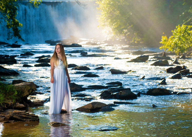 A picture of a girl who is a senior posing for her senior portrait wearing a long white dress as she stands on a river rock surrounded by a flowing river with a waterfall in the background as sunlight streams through the trees, Moreland Photography, Senior portrait, portrait, portraiture, long white dress, white dress, senior picture, senior, river, river rocks, waterfall, light, Creative lighting, lighting, sunlight, water, greenery, atlanta, atlanta scenery, atlanta waterfall, atlanta river