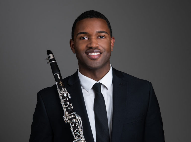 A man smiling for his business headshot while holding his instrument and wearing a black suit with a white dress shirt underneath and a black tie on, Moreland Photography, Atlanta headshot photography, business headshot, professional headshot, atlanta photographer, business headshot portrait, black suit, white dress shirt, instrument, black tie 