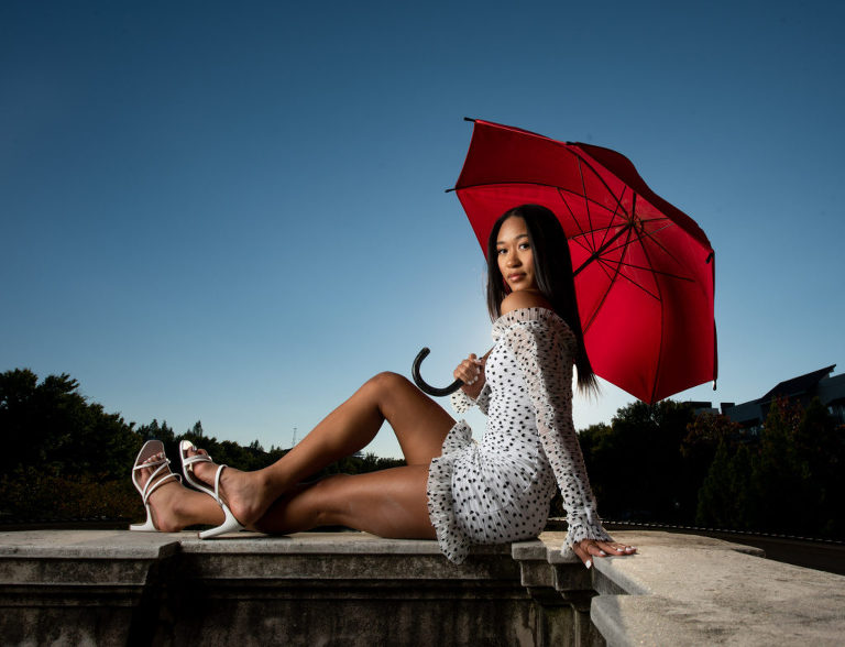 A girl wearing a white short lengthen dress with black polkadots and ruffles with matching white strap heels and matching white nail polish while also holding a vibrant red umbrella as she sits down, Moreland Photography, Senior Portrait, Portrait, Portraiture, Red Umbrella, Umbrella, Creative Lighting, Lighting, White Dress, Polka Dots, White dress with black polka dots, dress with ruffles, white heels, white nail polish, 