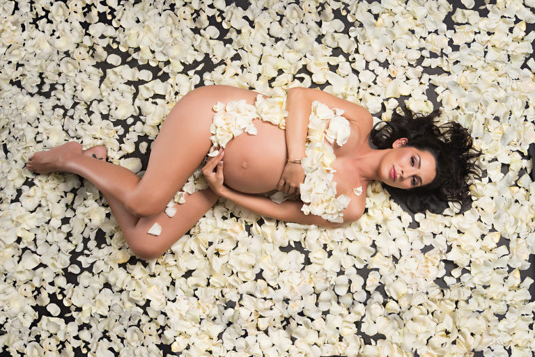 A pregnant woman smiling for her magazine style maternity photoshoot as she cradles her pregnant belly and is surrounded by white flower petals that are around her as well as on her, Moreland Photography, Atlanta maternity photography, magazine style maternity photo, maternity portrait,  Atlanta maternity portrait, Flower petals, white flower petals, flowers